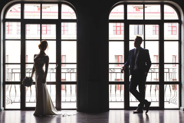 身着西装的男人和一个女人穿着裙子，在一个别致的空旷的室内一个大型全景彩色玻璃窗附近相映成轻的影子剪影 — 图库照片