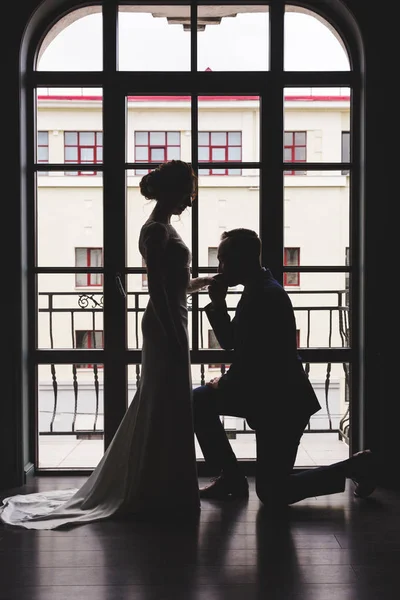 Silhouettes of a man in a suit and women in a dress and with a bouquet of flowers. The man knelt down in front of his lover, took her hand and kissed her near a large panoramic stained glass window. — Stock Photo, Image