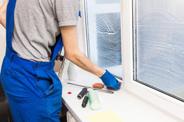 El primer plano de un hombre en uniforme y guantes azules lava las ventanas con rascador de ventanas. Servicio profesional de limpieza en el hogar — Foto de Stock