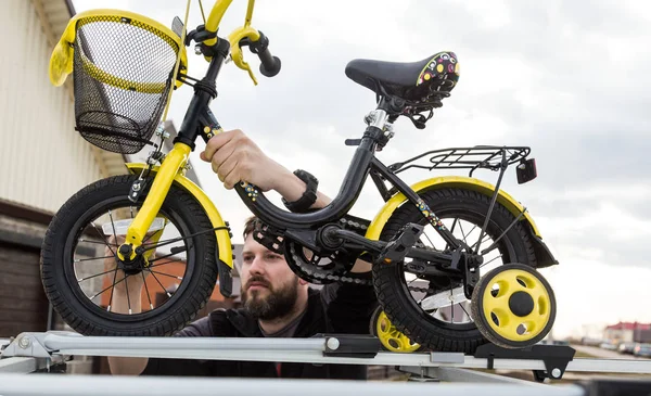 Fietstransport-een man bevestigt en installeert een Kinderfiets op het dak van een auto in een speciale houder voor fiets vervoer. De beslissing om grote ladingen te vervoeren en te reizen met de auto. — Stockfoto