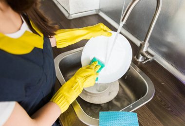 Sarı eldivenli genç bir kadın bulaşıkları lavaboda süngerle yıkıyor. Profesyonel ev temizleme servisi.