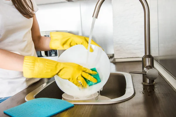 Een jonge vrouw met gele handschoenen wast de vaat met een spons in de gootsteen. Professionele schoonmaakdienst. — Stockfoto