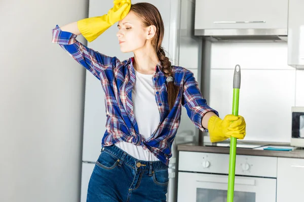 En ung kvinna städar hemma, tvättar köket. Skopa med trasor och gula handskar på bordet — Stockfoto