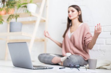 Genç, mutlu bir kadın yoga pozunda, parlak bir dairede oturuyor ve dizüstü bilgisayarın arkasında çalışıyor. Serbest çalışan bir kız çalışırken meditasyon yapıyor. İş stresi.