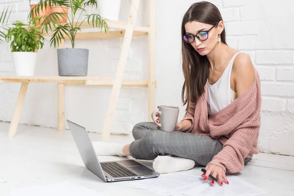 Mulher jovem senta-se no chão em um interior apartamento escandinavo com um laptop, estudando direito, menina freelance no trabalho, estudante de ensino à distância, emprego on-line, papelada e conceito de terceirização — Fotografia de Stock