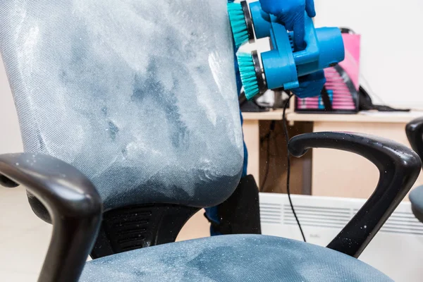 Giovane uomo in abbigliamento da lavoro e guanti di gomma pulisce la sedia dell'ufficio con attrezzature professionali . — Foto Stock