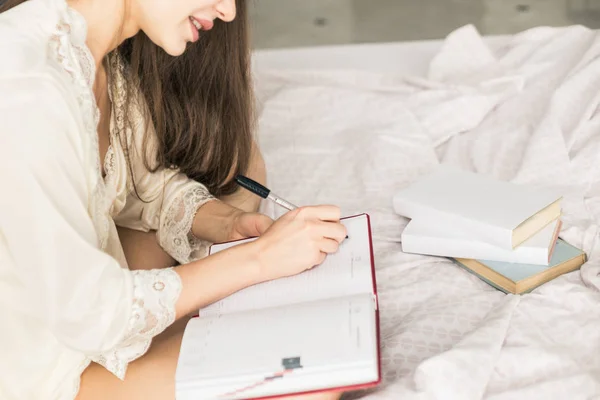 Aantrekkelijk jong meisje in een nachtjapon zit in bed en schrijft in een notitieboekje. — Stockfoto