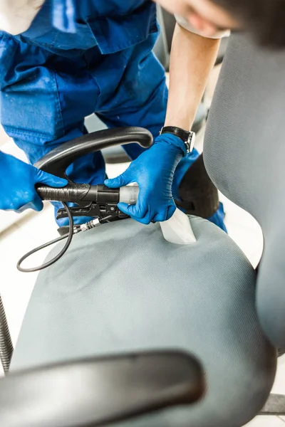 Hombre joven en ropa de trabajo y guantes de goma limpia la silla de la oficina con equipo profesional . — Foto de Stock