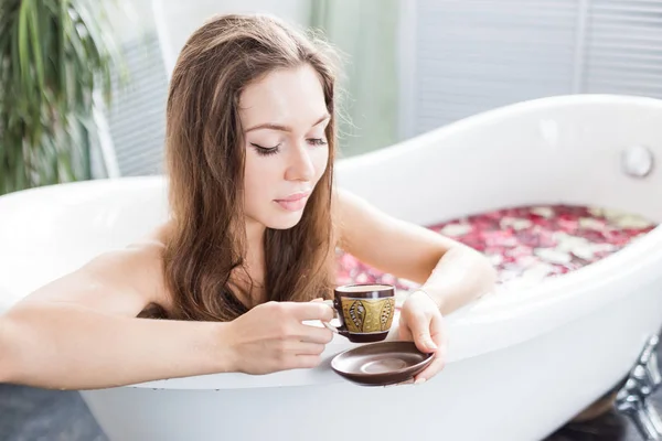 Una giovane ragazza attraente prende un bagno con petali di fiori e si rilassa sullo sfondo di un bellissimo interno leggero. Trattamenti Spa per la bellezza e la salute con la cura della pelle — Foto Stock