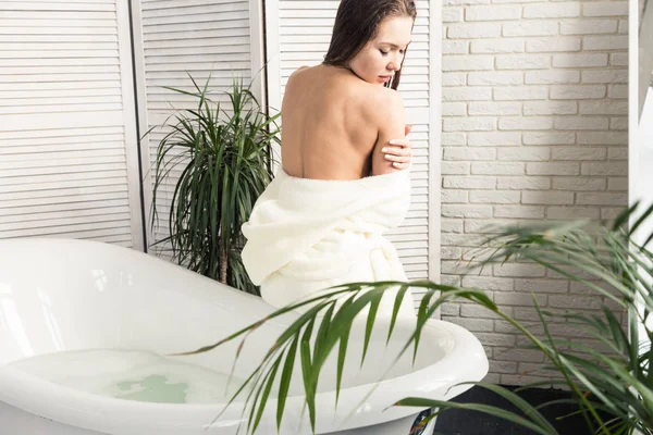 Atraktivní mladá dívka v bílém županu nanáší krém na kůži, zatímco sedí v koupelně a provádí ranní procedury. Péče o pleť a tělo — Stock fotografie