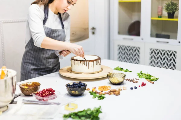 A confectioner squeezes liquid chocolate from a pastry bag onto a white cream biscuit cake on a wooden stand. The concept of homemade pastry, cooking cakes. — Stock Photo, Image