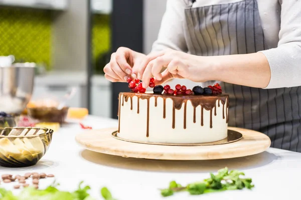 De banketbakkerij versiert met bessen een koekjes cake met witte room en chocolade. Cake staat op een houten stand op een witte tafel. Het concept van zelfgemaakte gebak, koken cakes. — Stockfoto