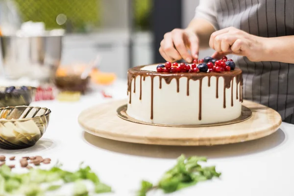 Confectioner menghiasi dengan beri kue biskuit dengan krim putih dan cokelat. Kue berdiri di sebuah berdiri kayu di atas meja putih. Konsep kue buatan sendiri, memasak kue . — Stok Foto