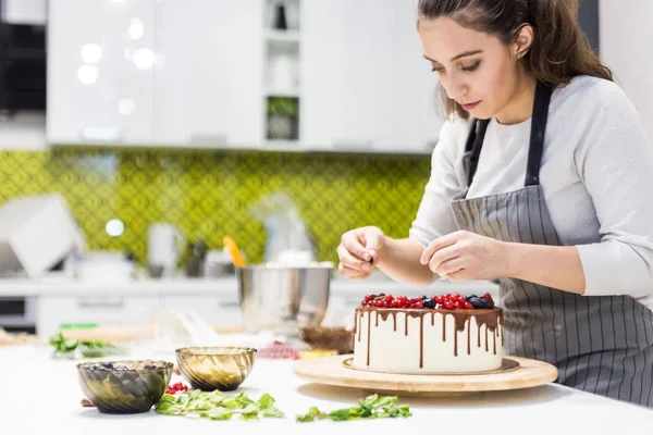 Cukrászda díszíti bogyókat keksz torta, fehér tejszín és csokoládé. A sütemény egy fából készült állványon áll egy fehér asztalon. A koncepció a házi készítésű péksütemények, főzősütemények. — Stock Fotó