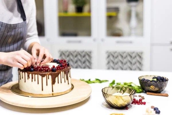 Confectioner dekorerar med bär en kak kaka med vit grädde och choklad. Kakan står på ett trä stativ på ett vitt bord. Konceptet med hembakat bak verk, matlagnings tårtor. — Stockfoto