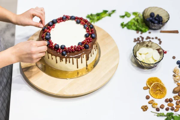 Confectioner decorates with berries a biscuit cake with white cream and chocolate. Cake stands on a wooden stand on a white table. The concept of homemade pastry, cooking cakes. — Stock Photo, Image