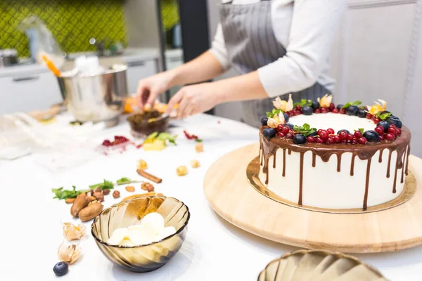 Il pasticcere decora con bacche una torta di biscotto con crema bianca e cioccolato. Cake si trova su un supporto di legno su un tavolo bianco. Il concetto di pasticceria fatta in casa, torte di cottura . — Foto Stock