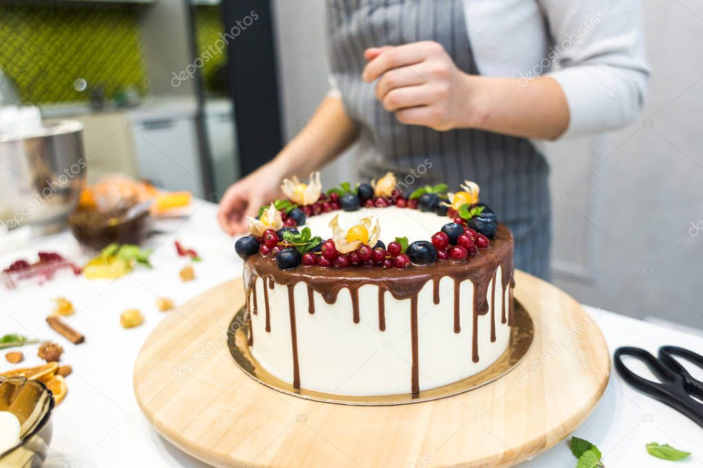 Confectioner decorates with berries a biscuit cake with white cream and chocolate. Cake stands on a wooden stand on a white table. The concept of homemade pastry, cooking cakes.