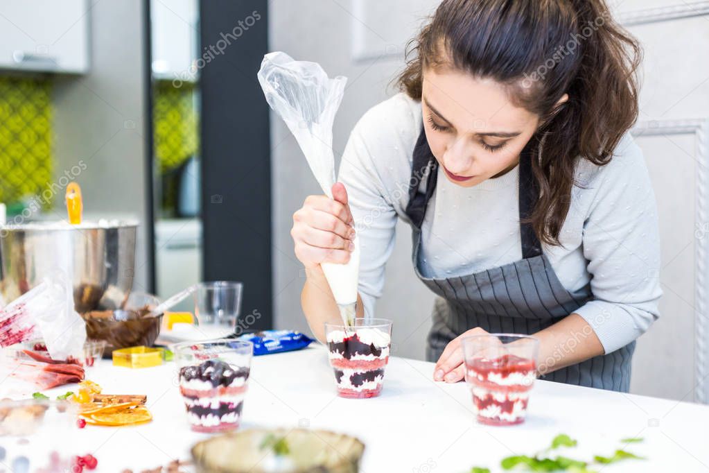 A confectioner prepares a trifle in three cups. Desserts are on the white table in the kitchen. The concept of homemade pastry, cooking cakes.