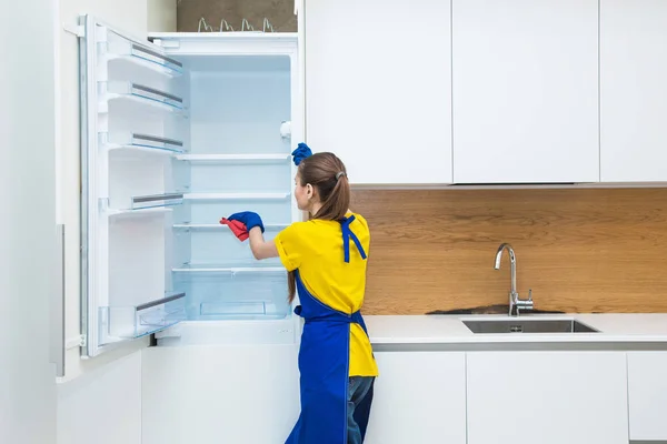 Servicio de limpieza profesional. Dos mujeres en uniforme de trabajo, en delantales, dividen la limpieza de la cocina de una casa privada, casa de campo. lavar el refrigerador, grifo, fregadero. Lavar el suelo. —  Fotos de Stock