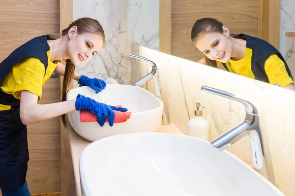 Profesjonalne sprzątanie. Kobieta w mundurze i rękawiczki gąbka myje kanalizację w łazience. Myjnia i umywalka — Zdjęcie stockowe
