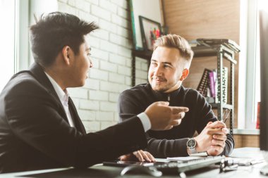 Masada oturan genç bir ofis çalışanı, bilgisayar kullanıyor. İki iş adamı konuşuyor.
