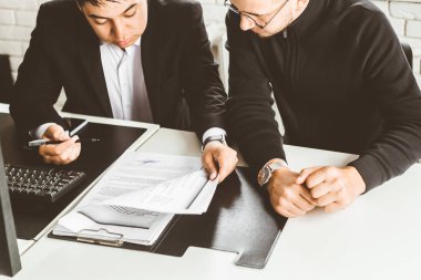 Masada oturan genç bir ofis çalışanı, bilgisayar kullanıyor. İki iş adamı konuşuyor.