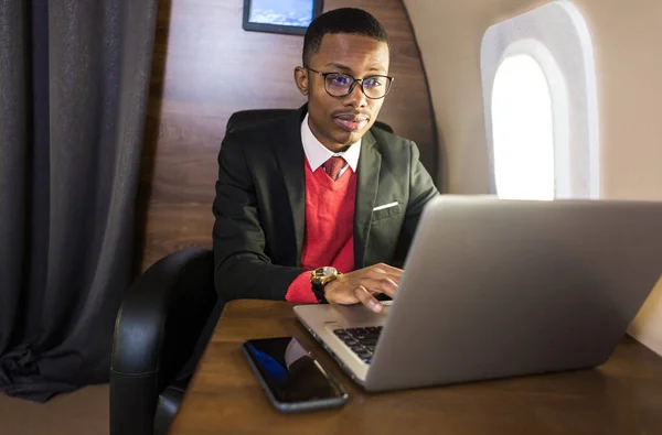 Atrakcyjny i udany African American biznesmen z okularami pracujących na laptopie siedząc na krześle jego prywatnego odrzutowca — Zdjęcie stockowe