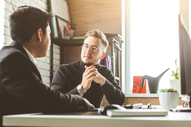 Masada oturan genç bir ofis çalışanı, bilgisayar kullanıyor. İki iş adamı konuşuyor.