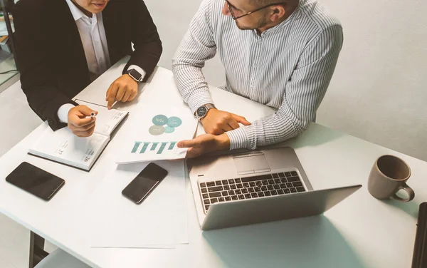 Masada oturan genç bir ofis çalışanı, bilgisayar kullanıyor. İki iş adamı konuşuyor.