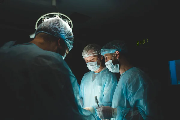 An international professional team of surgeon, assistants and anesthesiologist perform a complex operation on a patient under general anesthesia. Dark atmospheric photography theme in low key.