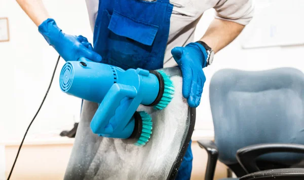 Young man in workwear and rubber gloves cleans the office chair with professional equipment. — Stock Photo, Image