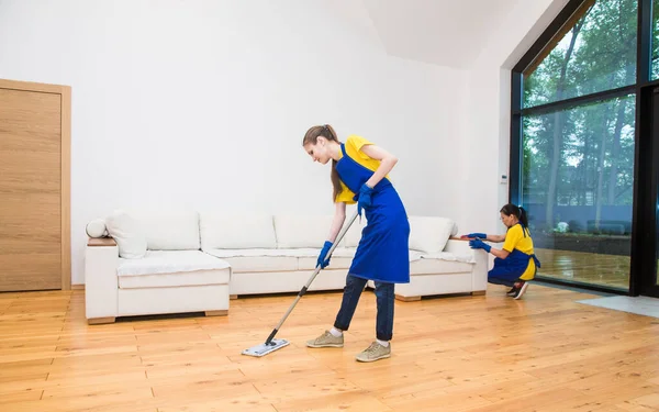 Profesyonel temizlik servisi. Önlüklü, işçi üniformalı iki kadın özel bir evin mutfağının temizliğini bölüşüyor. Yerleri siliyorum.. — Stok fotoğraf
