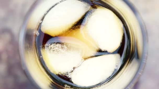 Κοντινό πλάνο ενός ποτηριού με ένα δροσιστικό κρύο ποτό με κομμάτια πάγου και υγρό φυσαλίδων σε θολή καλοκαιρινή φυλλωσιά — Αρχείο Βίντεο
