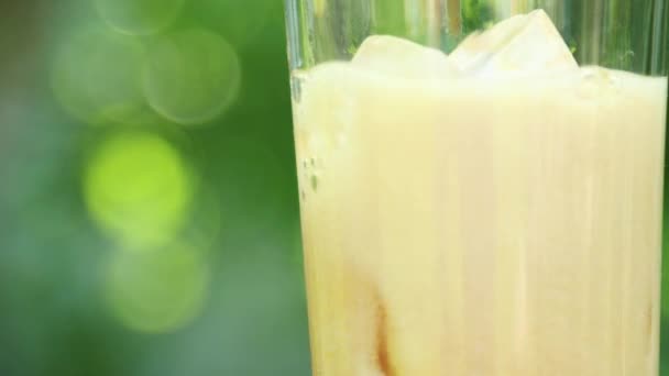 Close-up of a glass with a refreshing cold drink with pieces of ice and bubbling liquid on a blurry summer foliage background — Stock Video