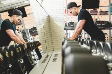 Atletik adam spor salonunda ayna önünde dambıllarla antrenman yapıyor.