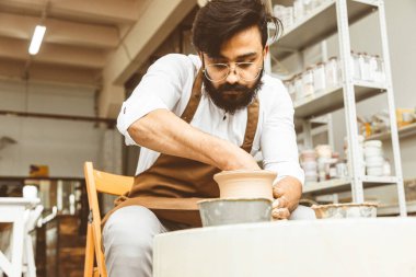 Genç bir erkek çömlekçi atölyesinde çömlekçi çarkında el işi yapıyor ve kilden bir ürün yapıyor.