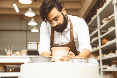 Genç bir erkek çömlekçi atölyesinde çömlekçi çarkında el işi yapıyor ve kilden bir ürün yapıyor.