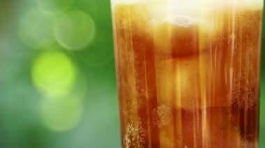 Bulanık bir yaz yeşilliği arka planında buzlu ve köpüren sıvıyla ferahlatıcı soğuk bir içecek içeren bir bardağın yakın çekimi.