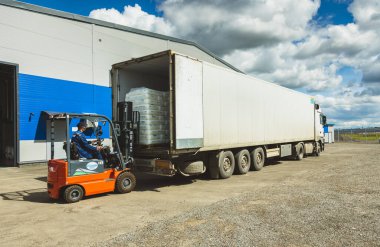 Forklift kullanan bir adam büyük bir depoda çalışır ve nakliye için bir kamyona hammadde yükler.