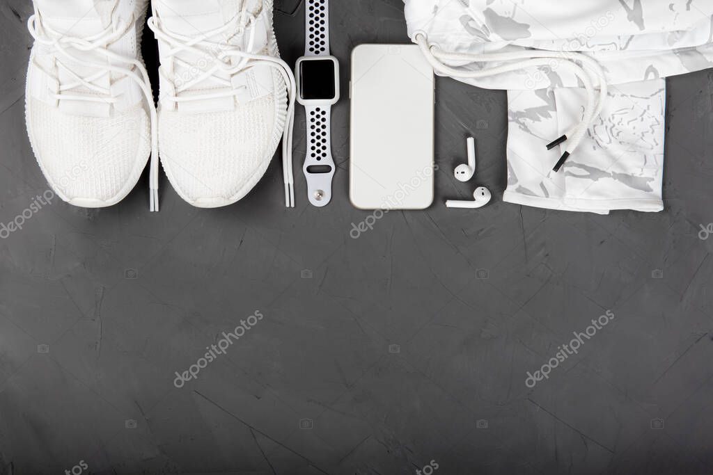 Fit and healthy lifestyle concept. Stylish fitness accessories: sneakers, camouflage training hoodie, smartphone, smartwatch, wireless earphones on dark gray concrete background. Flat lay, copy space.