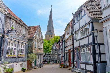 Hattingen, Almanya - 14 Haziran 2025: Geleneksel yarı keresteli evler ve Almanya 'nın Hattingen kentinde açık mavi bir gökyüzü altında önde gelen bir kilise kulesi ile sıralanmış büyüleyici kaldırım taşı sokakları.