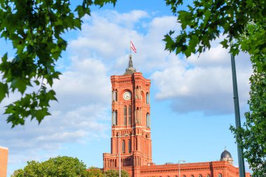 Berlin, Almanya - 17 Ağustos 2025: Tarihi Red City Hall Kulesi, yeşil yapraklar çerçeveli, tüylü, mavi bir gökyüzüne karşı yükseliyor.