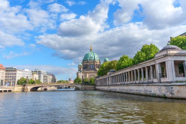 Berlin, Almanya - 17 Ağustos 2025: Geniş bulutlu bir gökyüzü altında Spree Nehri boyunca ağaçlar tarafından çerçevelenmiş yeşil kubbeli Berlin Katedrali.