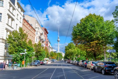 Berlin, Almanya - 17 Ağustos 2025: Ağaçlarla kaplı bir cadde boyunca uzanan tramvay rayları ve arkadaki Berlin TV Kulesi parçalı bulutlu bir gökyüzünün altında.
