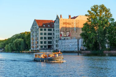 Berlin, Almanya - 18 Ağustos 2025: Berlin 'deki Spree Nehri' nde süzülen tekne, gün batımında tarihi binalar ve şehir grafitileri sergiliyor..