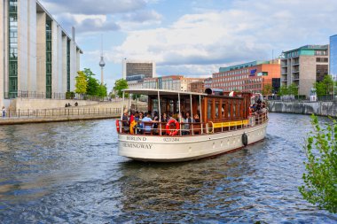 Berlin, Almanya - 20 Ağustos 2025: Hemingway adlı zarif nehir teknesi Spree Nehri boyunca süzülüyor, çağdaş binalar ve parçalı bulutlu bir gökyüzü altında yemyeşil alanlar tarafından çevreleniyor..