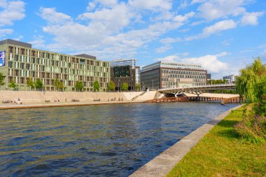 Berlin, Almanya - 20 Ağustos 2025: Çağdaş binalar Spree Nehri 'ni yansıtırken, insanlar mavi gökyüzü altında dağınık bulutlarla dinleniyorlar..