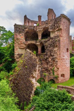 Historic stone ruins of Heidelberg Castle with crumbling walls, lush foliage, and dramatic cloud formations in the background.