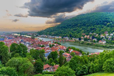 Günbatımında dramatik bir gökyüzü altında tarihi Heidelberg, Neckar Nehri ve yemyeşil tepeleri kaplayan panoramik manzara.